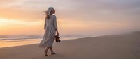 Woman in light linen dress walking barefoot on sandy beach at golden hour sunset, holding shoes in hand, peaceful coastal scene.
