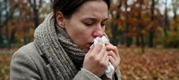 Woman with tissue suffering from cold or flu symptoms in autumn park setting with warm knitted scarf and sweater for healthcare and seasonal illness concepts.