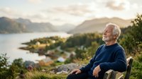 Senior man with white hair sitting outdoors overlooking mountain lake and forested valley landscape during golden hour, contemplative lifestyle portrait.
