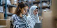 Young woman with long dark hair and South Asian ethnicity wearing striped shirt working at computer in office with colleague in hijab in background, teamwork and professional environment.