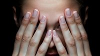 Woman covering face with both hands showing french manicured nails against dark background, expressing emotion of fear, shame, or hiding from problems.
