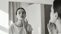 Caucasian woman in casual white t-shirt examining her face in mirror, touching chin thoughtfully in bright bathroom with natural sunlight. Skincare, self-care routine.