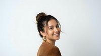 Young Hispanic woman with curly hair in bun wearing brown sweater smiling at camera against white background for portrait photography and lifestyle content.