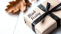 Black Friday gift box with elegant black ribbon and tag on white wooden background with autumn leaves for holiday shopping promotions and seasonal sales campaigns.