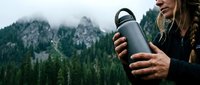 Young Caucasian woman hiker holding stainless steel water bottle in mountain wilderness with forest and rocky peaks background for outdoor adventure lifestyle.