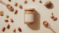 Creamy almond butter in glass jar surrounded by scattered whole almonds, walnuts and wooden spoon on beige background. Natural food photography layout.