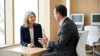 Two business professionals, a middle-aged Caucasian woman and man in suits, having a friendly meeting discussion in a modern office.