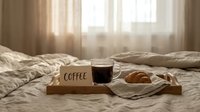 Cozy morning breakfast tray on bed with black coffee in glass cup, croissant, and coffee sign in soft natural light bedroom.