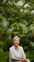 Senior woman with gray hair and East Asian ethnicity smiling in lush garden surrounded by green foliage and plants, wearing light beige shirt, outdoor portrait for wellness and lifestyle content.