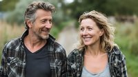 Mature Caucasian couple smiling at each other on rural path, wearing casual plaid shirts, surrounded by natural greenery in outdoor setting.