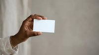 Man with dark skin tone holding blank white business card or invitation in hand against neutral background for branding mockup.