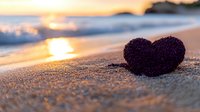 Purple heart shape on sandy beach at golden sunset with ocean waves and glowing sun reflection, romantic seaside scene for Valentine's Day cards and love concepts.