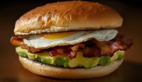 Appetizing breakfast sandwich with crispy bacon, fried egg, fresh avocado slices on toasted bun against dark background, close up view of morning meal.