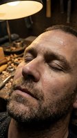 Middle-aged man with beard and closed eyes resting under warm desk lamp in home office setting. Caucasian male appearing relaxed or fatigued during work.