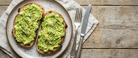 Fresh avocado toast on rustic sourdough bread served on ceramic plate with golden fork and knife on wooden table background for healthy breakfast menu.