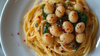 Fresh spaghetti pasta with scallops, spinach and red pepper flakes served on white plate, close up view of traditional Italian seafood dish.