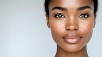Young African American woman with glowing natural makeup and flawless skin against light background, expressing confidence and beauty in minimalist portrait.