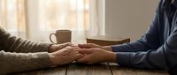 Elderly couple holding hands across wooden table with coffee mug in warm sunlit room showing love, support and companionship in golden years.