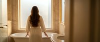Young woman in white robe standing by luxury bathtub in bright bathroom with natural light from windows, spa wellness relaxation morning routine.