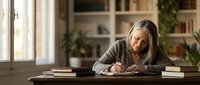 Senior woman with gray hair writing in notebook at wooden desk in bright home office with bookshelves and plants, focused on personal journaling or work.