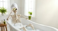 Human skeleton relaxing in bathtub with towel on head holding green smoothie in bright bathroom with plants for humorous health and wellness concept.