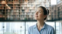 Woman in business casual attire gazing upward thoughtfully near office windows with urban cityscape visible behind glass, conveying aspiration and professional contemplation.