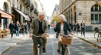 Senior couple riding vintage bicycles together on charming European city street with historic architecture and pedestrians in background.