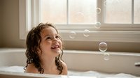 Young girl with curly hair enjoying bubble bath time in white bathtub by window with natural light creating joyful childhood moment for family lifestyle content.