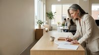 Mature woman with long gray hair and glasses, wearing a beige cardigan, writing or taking notes at a wooden desk in a bright, modern office space with natural light and potted plants.