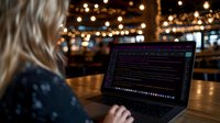 Blonde woman programmer coding on laptop in cozy cafe with warm string lights creating productive remote work atmosphere for tech professionals.