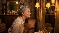 Senior Caucasian woman with gray hair smiling while looking in ornate vintage mirror in elegant room with warm lighting and classic decor.