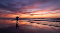 Silhouette of angler fishing at beach during dramatic sunset with purple and orange sky reflecting on wet sand and ocean waves.