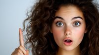 Young woman with curly dark hair and surprised expression raising index finger in eureka moment against light background. Concept of idea or solution.