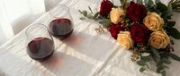 Romantic evening setup with red wine glasses and mixed rose bouquet on white linen tablecloth for Valentine's Day celebration or anniversary dinner.