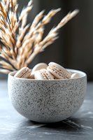 French macarons in speckled ceramic bowl with dried pampas grass decoration against dark background. Minimalist food styling for cafe and bakery marketing.