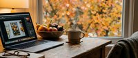 Cozy autumn workspace with laptop, steaming coffee mug, fruit bowl, and glasses on rustic wooden desk by window with fall foliage.