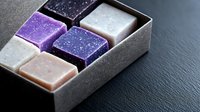 Handcrafted soap bars in purple, pink, white and black colors with glitter effect arranged in gray box on dark background. Natural skincare and bath products.