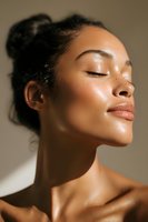 Young African American woman with glowing skin and closed eyes enjoying sunlight, natural makeup look and hair in bun against neutral background.