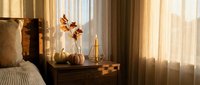 Cozy bedroom interior with warm sunlight streaming through sheer curtains onto wooden nightstand with decorative vase and autumn branches creating peaceful atmosphere.