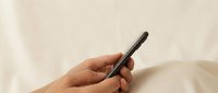 Hand holding black smartphone with blank screen in bed with white sheets for mobile technology and communication concepts.