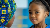 Young African girl with braided hair smiling warmly while looking at another person wearing traditional blue and yellow patterned headwrap, against blurred blue background.