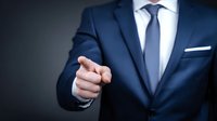 Businessman in navy blue suit pointing finger forward against dark background, conveying leadership and confidence in professional setting.