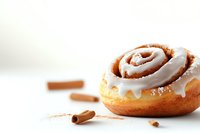 Fresh baked cinnamon roll with white cream cheese frosting and cinnamon sticks on light background, selective focus on sweet pastry dessert.