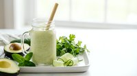 Green smoothie with avocado and cucumber in glass jar with bamboo straw on white tray, fresh herbs and sliced vegetables for healthy breakfast.