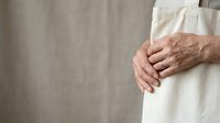 Senior adult male with weathered hands wearing cream linen clothing, holding fabric against neutral beige wall. Suitable for healthcare, aging, wellness, and lifestyle editorial use.