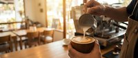 Professional barista pouring steamed milk creating latte art in coffee cup at modern cafe with warm lighting and blurred restaurant background.