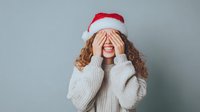 Young Caucasian woman with curly hair wearing Santa hat and cozy knit sweater covering eyes with hands against neutral gray background. Holiday anticipation concept.