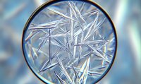 Microscopic view of crystalline ice formations showing delicate needle-like structures in blue tones against blurred background, captured through circular lens.