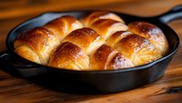 Fresh baked golden brown dinner rolls in cast iron skillet with glossy buttery crust, showing soft texture and braided pattern on rustic wooden surface.