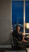 Thoughtful businesswoman relaxing in modern apartment chair by floor-to-ceiling windows overlooking illuminated city skyline at night.